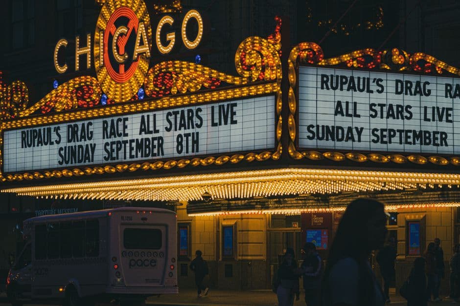 The lit marquee of the Chicago Theatre at night with the word Chicago in bulbs above it, people walking past on the sidewalk and a small white bus at the curb