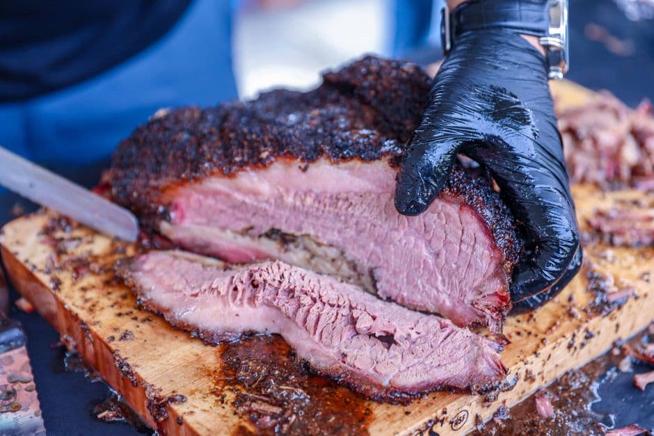 Gloved hands slicing a whole smoked brisket with a long knife on a wooden board, showing the bark and smoke ring