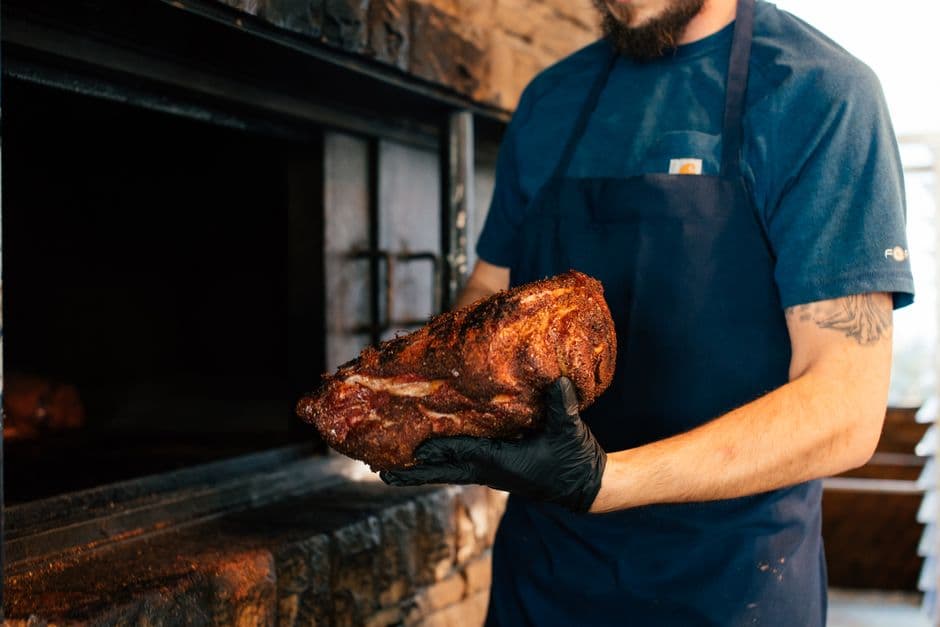 A pitmaster in an apron and black glove lifting a smoked brisket out of a brick barbecue pit