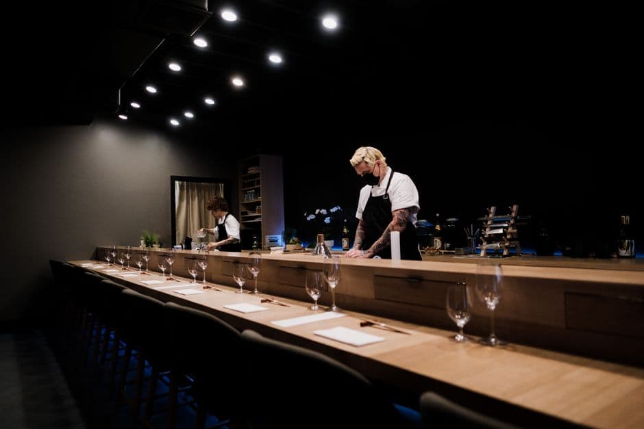 A long wooden omakase counter set with wine glasses and napkins while two chefs prepare service in a dark dining room