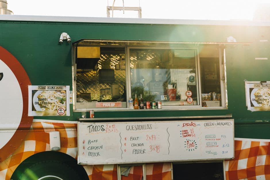A food truck service window with a hand lettered menu board listing tacos, quesadillas, chilaquiles and drinks with prices