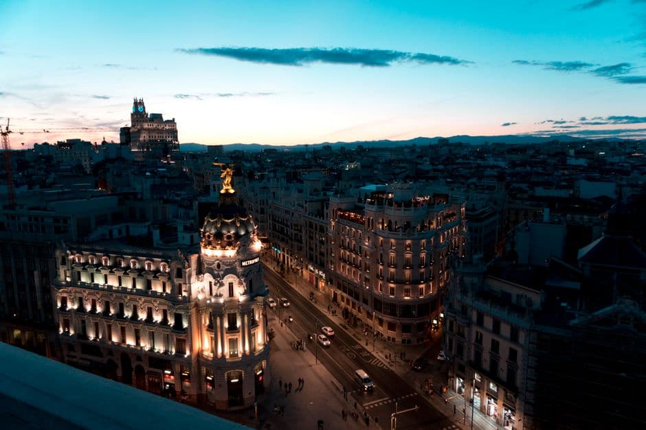 Gran Vía in Madrid at dusk seen from a rooftop, with the lit dome and winged statue of the Metropolis building in the foreground and the city stretching to the mountains