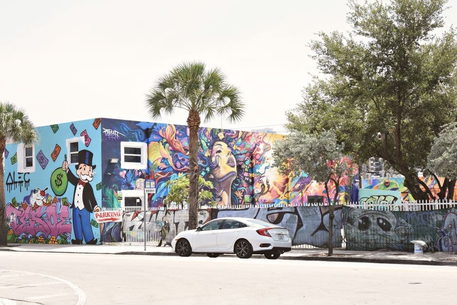 Colorful murals covering a low building and fence in Miami's Wynwood neighborhood, with palm trees and a parked car