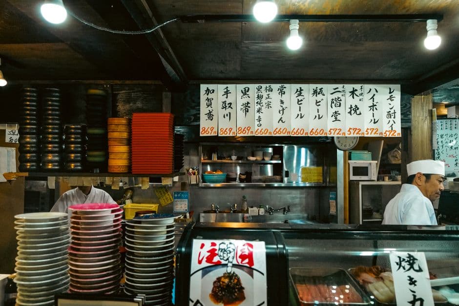 An everyday Japanese sushi counter with stacks of colored plates, a chef in white and a handwritten board of sake and beer prices in yen