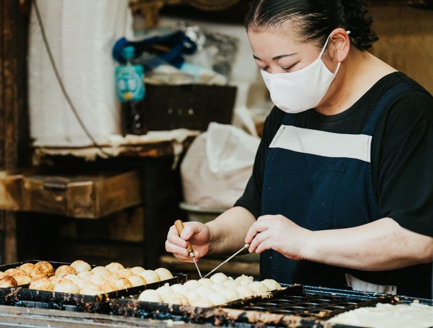A street vendor turning takoyaki balls in a cast-iron griddle with two thin picks