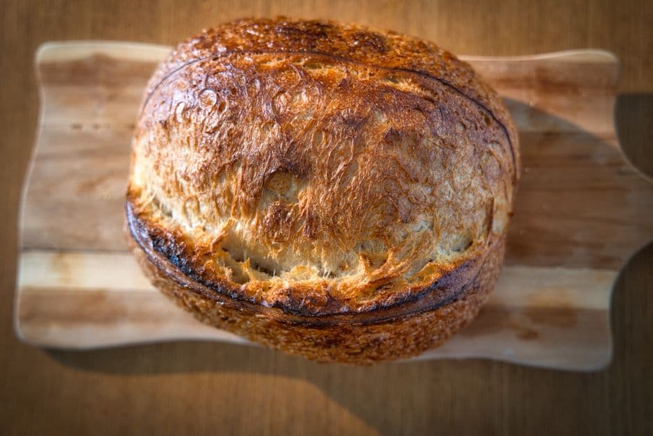 A round sourdough loaf on a wooden board with a dark blistered crust and a raised ear along the score