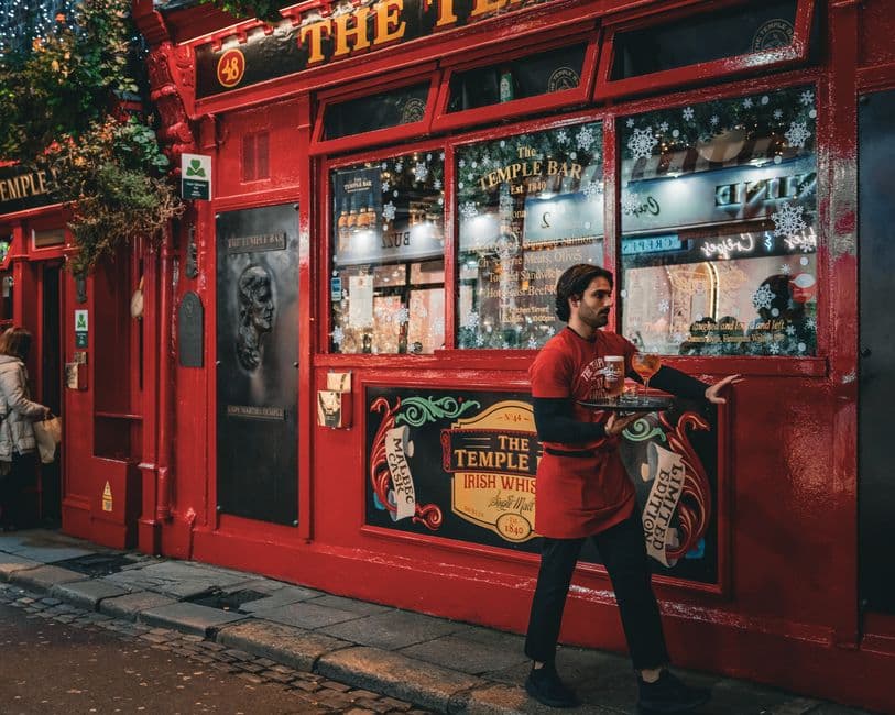 A waiter in a red apron carrying drinks on a tray past the red-painted front of The Temple Bar pub in Dublin at night, with Christmas decorations in the windows