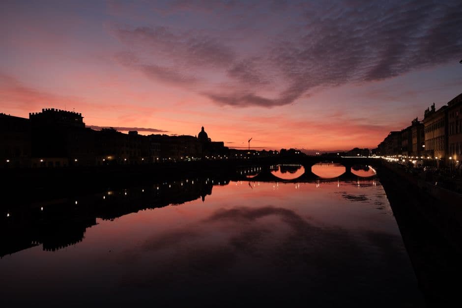 An arched stone bridge silhouetted across the Arno in Florence at sunset, with a pink and orange sky reflected in flat water and a domed church on the skyline