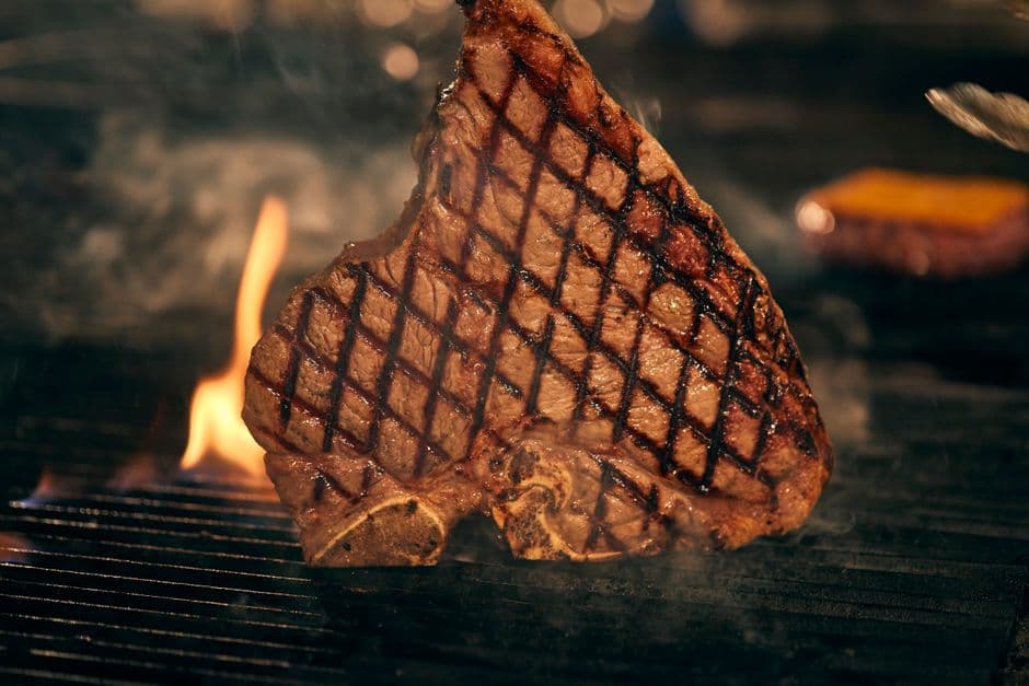 A thick T-bone steak searing on a grill over an open flame, with heavy cross-hatched grill marks and smoke rising around it