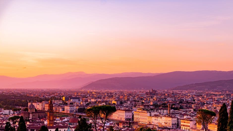 Florence seen from above at sunset, terracotta rooftops and church towers running to the Arno with the Tuscan hills and a pink and orange sky behind