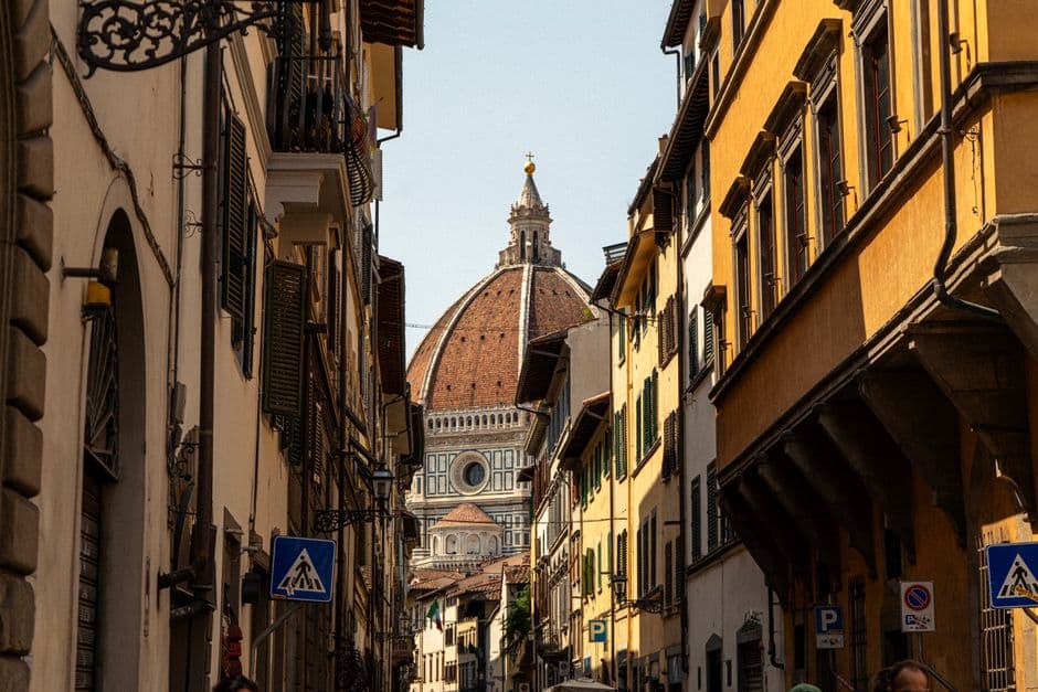 A narrow Florence street lined with shuttered ochre and cream buildings, with the terracotta dome and lantern of the cathedral filling the gap at the end