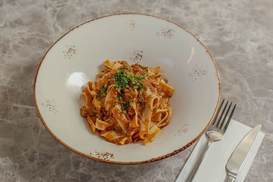 A shallow white bowl of flat ribbon pasta coated in a thick meat ragu and scattered with chopped herbs, on a pale marble surface next to a fork and knife