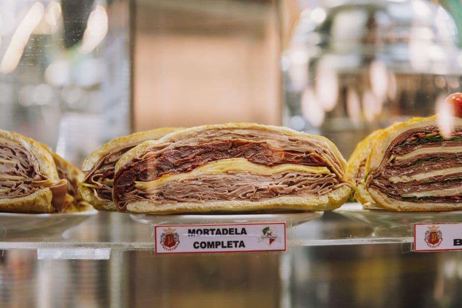 Three halved layered sandwiches on plates behind a glass deli counter, thick with folded cured meat and cheese, one labeled mortadela completa