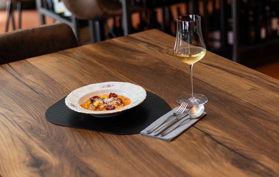 A single bowl of pasta in tomato sauce on a slate mat on a wooden restaurant table, with a glass of white wine and cutlery folded in a napkin beside it