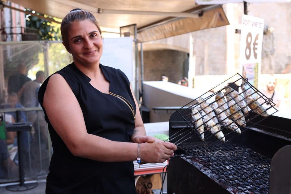 A cook turning a hinged wire basket of salt-crusted sardines over charcoal at a street grill, beside a sign advertising a port wine tasting for 8 euros