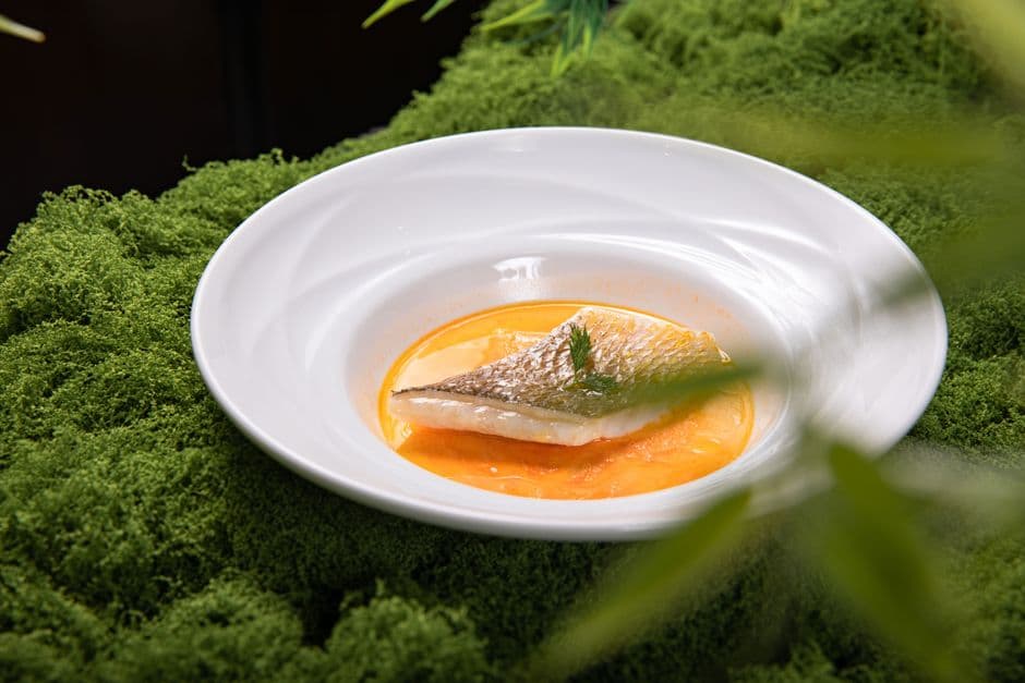 A fillet of white fish plated in an orange shellfish sauce in a shallow white bowl, set on green moss