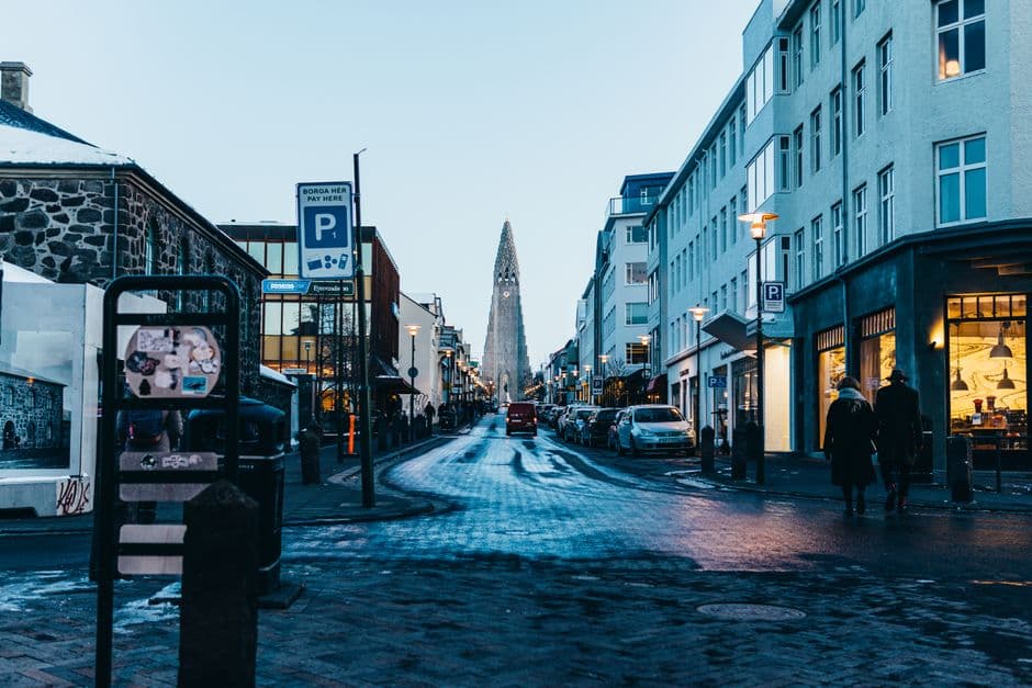 A wet cobbled street in central Reykjavik at dusk with lit shopfronts and the spire of Hallgrimskirkja at the far end