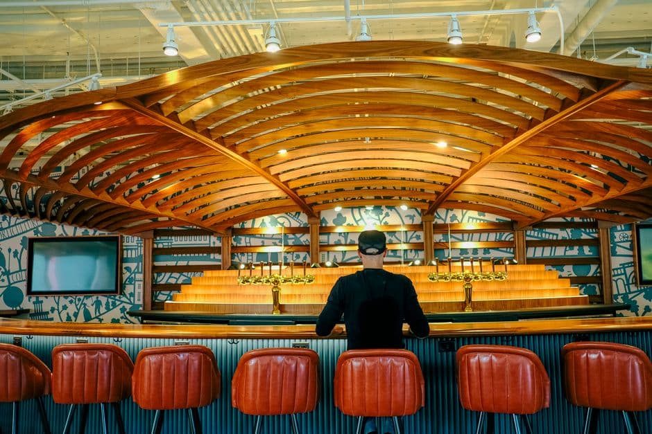 A person sitting alone at a long wooden bar counter on a red leather stool, under a curved timber ceiling, facing a row of brass taps