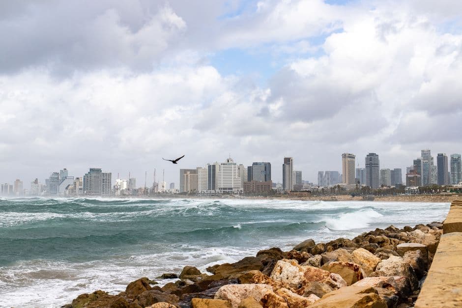 The Tel Aviv skyline seen across the sea from a rocky breakwater, with waves and a cloudy sky