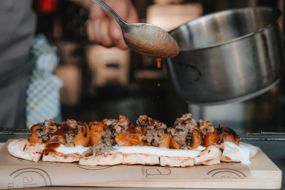 A cook spooning sauce over grilled meat and vegetables on flatbread at a street food counter