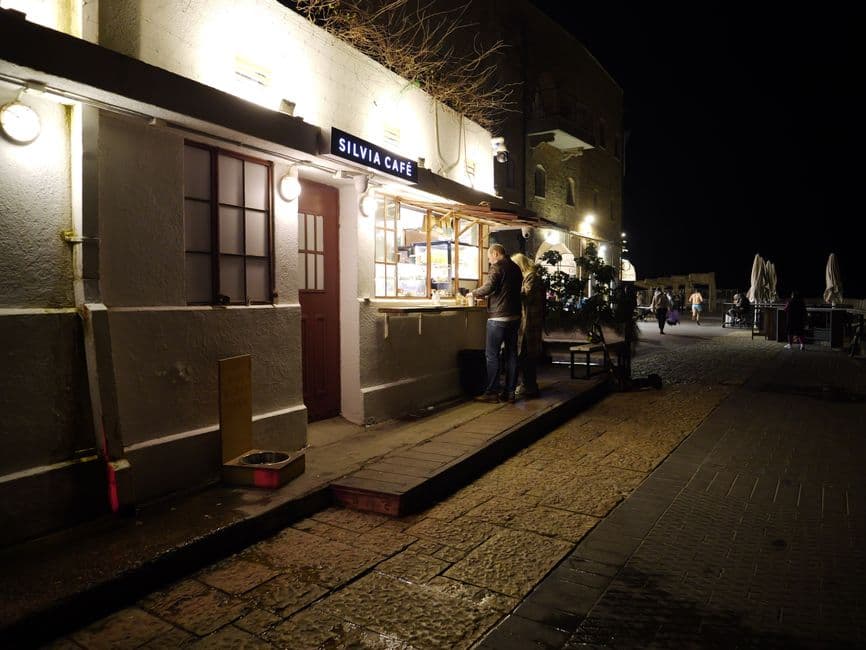 Silvia Cafe lit up at night with people ordering at a window onto a stone promenade
