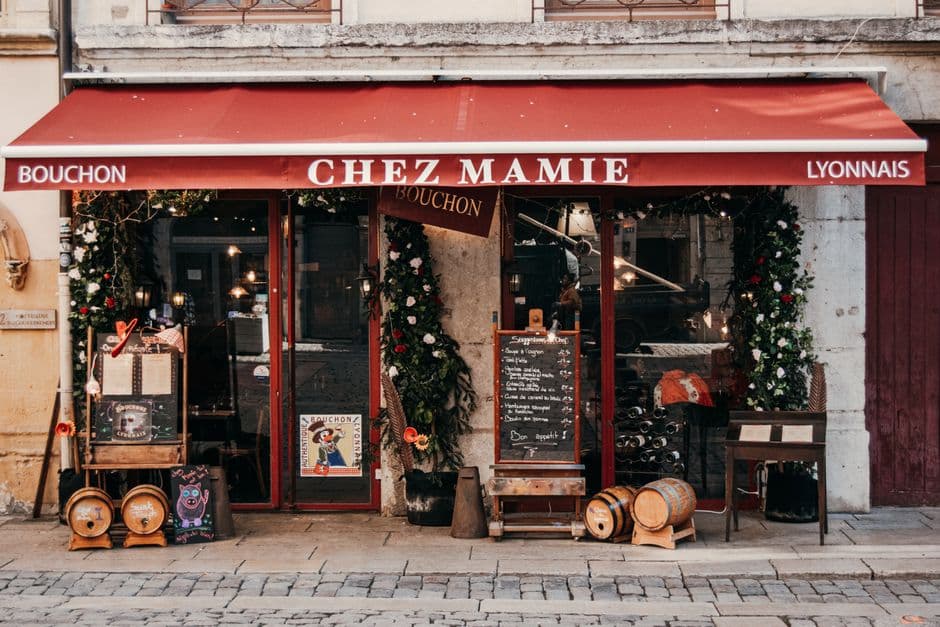 The front of Chez Mamie, a bouchon in Lyon, under a red awning reading Bouchon Chez Mamie Lyonnais, with a handwritten chalkboard menu, wine barrels on the cobbled sidewalk and an Authentique Bouchon Lyonnais poster in the door