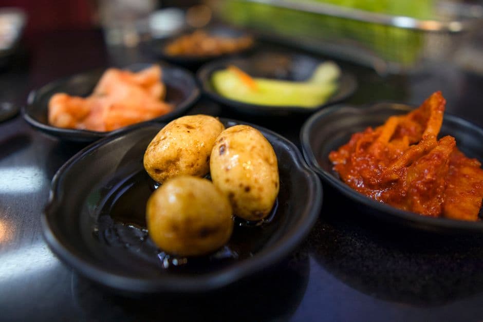 Six small black lacquer dishes of Korean banchan on a restaurant table, including napa cabbage kimchi, braised baby potatoes in soy, radish kimchi and sliced cucumber and pepper