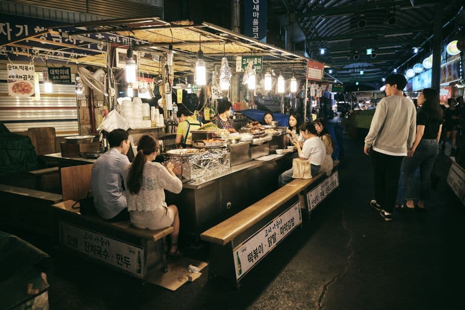 A covered Seoul market food alley at night, with stalls signposted in Korean for tteokbokki, sundae, mandu, janchi-guksu and mayak gimbap, and diners eating on benches at the stainless steel counters