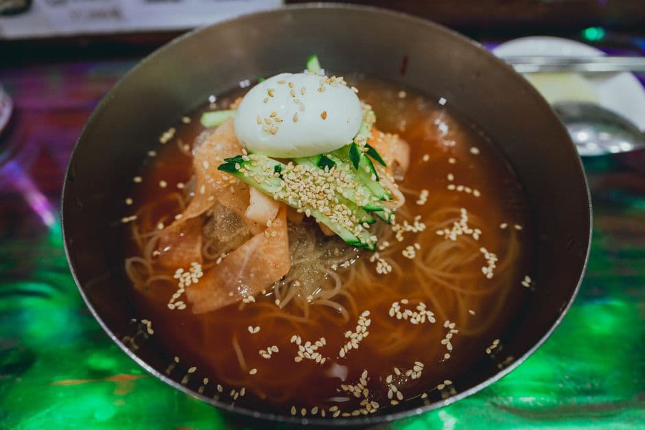 Cold noodles in a chilled brown broth served in the wide metal bowl Korean naengmyeon houses use, topped with half a boiled egg, sliced cucumber, pickled radish and sesame seeds