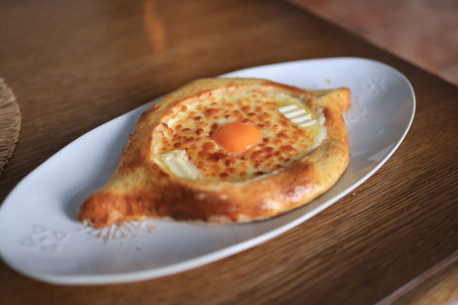 A boat-shaped Adjaruli khachapuri on a white oval plate, filled with browned melted cheese, a raw egg yolk in the center and two pats of butter