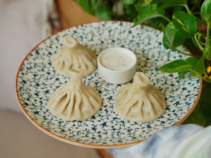 Three khinkali dumplings with pleated sides and twisted top knots on a blue and white patterned plate, with a small bowl of white sauce in the middle