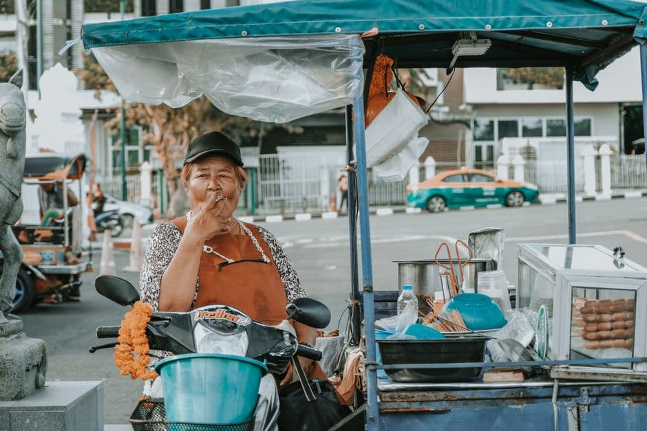 A Bangkok street food vendor sitting with her cart of utensils and snacks beside a parked motorbike, with a tuk-tuk and a green and orange Bangkok taxi in the background