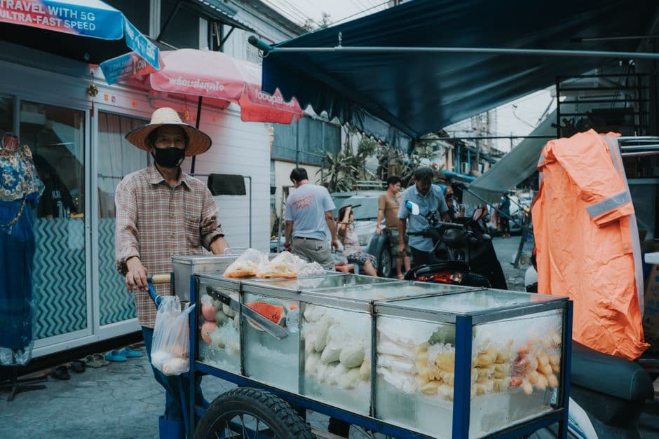 A Bangkok street vendor in a straw hat pushing a glass-cased cart of cut fruit on ice along a busy street