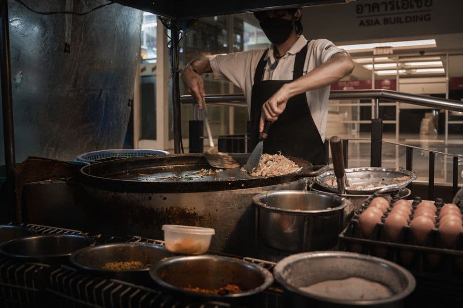 A cook stir-frying pad thai noodles in a wok on a street cart in Bangkok, with egg trays and condiment bowls lined up along the front