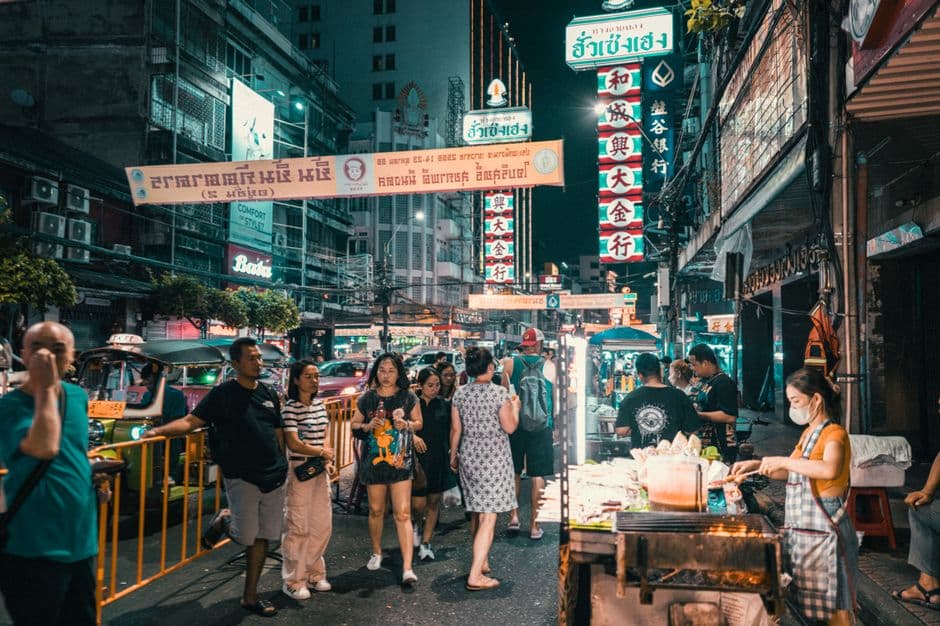 Crowds walking down Yaowarat Road in Bangkok's Chinatown at night under neon signs, with a grill cart in the foreground and a tuk-tuk behind