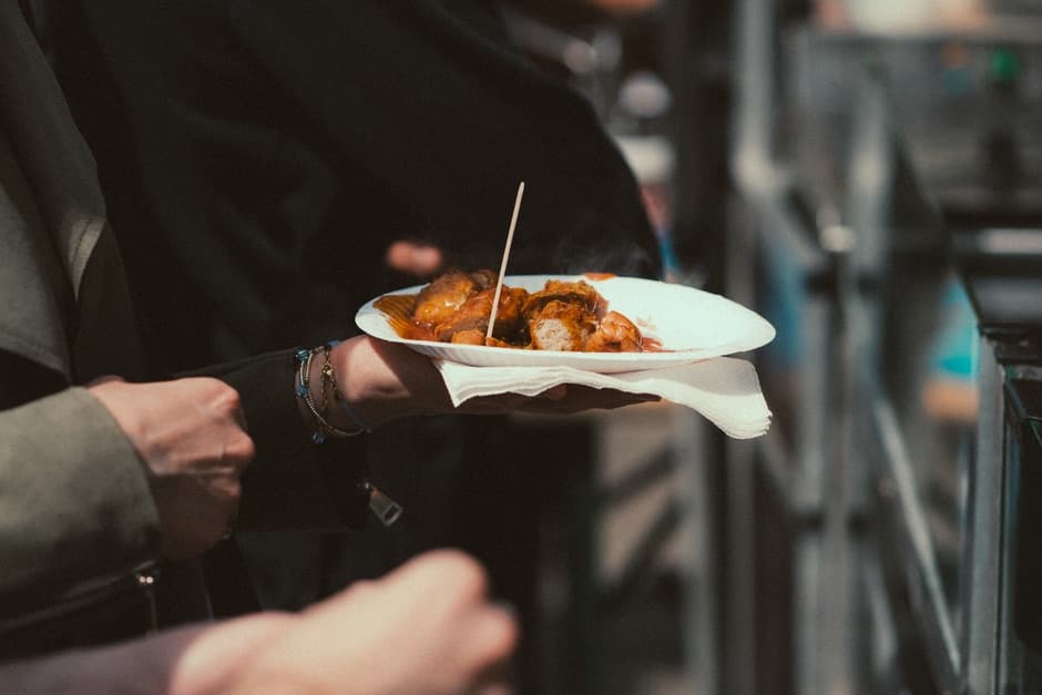 A hand holding a paper plate of sliced sausage in red curry sauce with a wooden pick, at an outdoor food stand