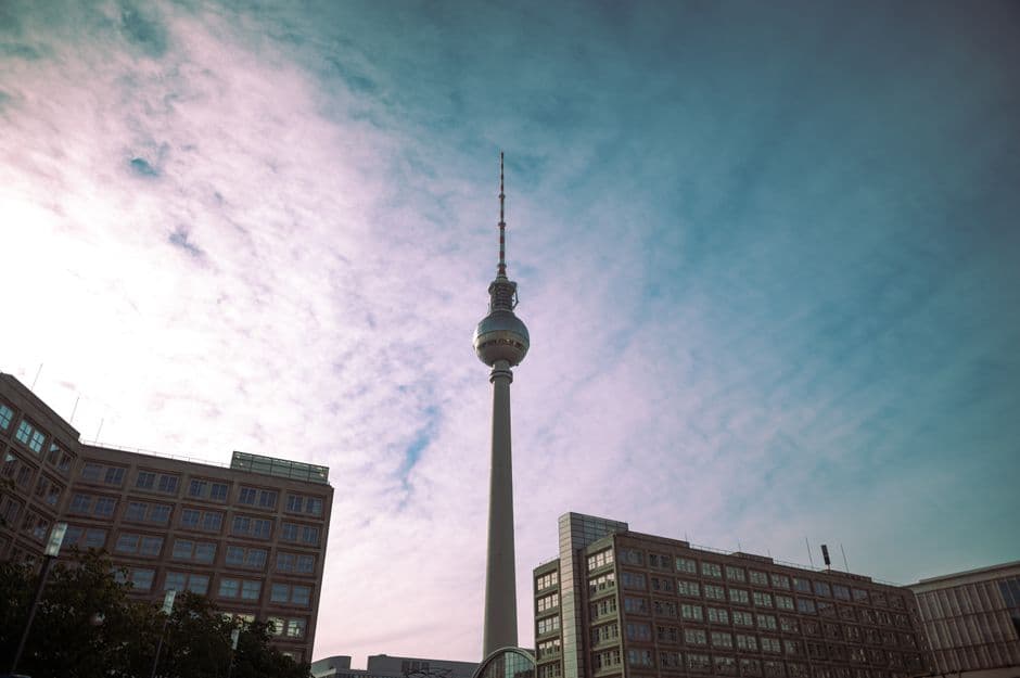 The Berlin TV Tower rising above office buildings at Alexanderplatz, seen from below against a pale blue and pink sky