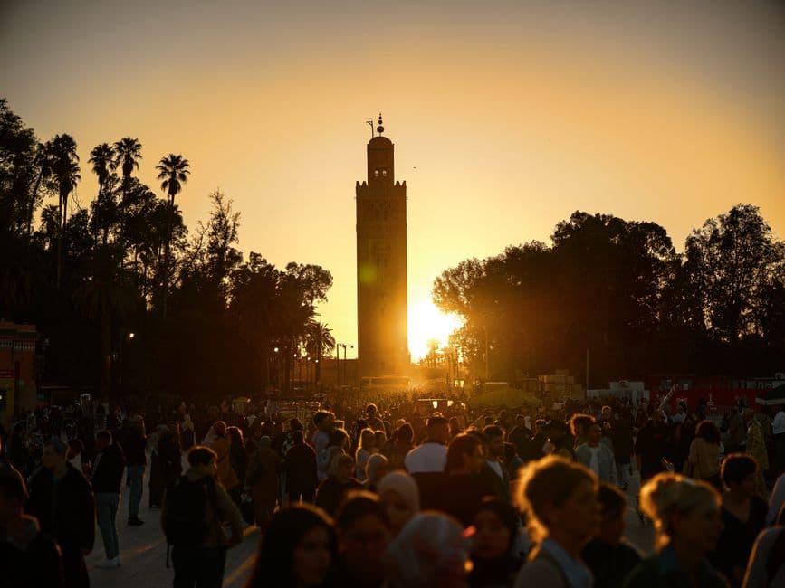 A crowd in silhouette in front of the Koutoubia minaret in Marrakech at sunset, with palm trees on either side under an orange sky
