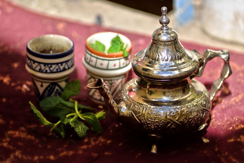 A silver Moroccan teapot beside two painted ceramic cups and a sprig of fresh mint on a red cloth