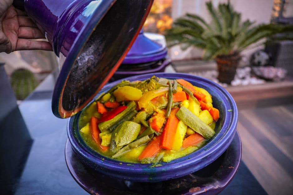 A hand lifting the conical lid of a blue glazed tagine to show a stew of carrots, zucchini, potatoes and green beans