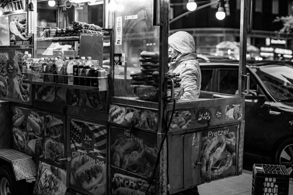 A black and white photograph of a New York street cart vendor with signs for chili cheese dogs, hot pretzels and Italian sausage