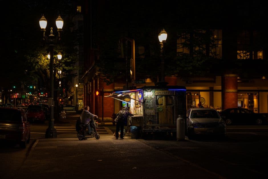 A food cart serving Mexican food lit up at night on a downtown street corner in Portland, Oregon, with twin globe streetlamps