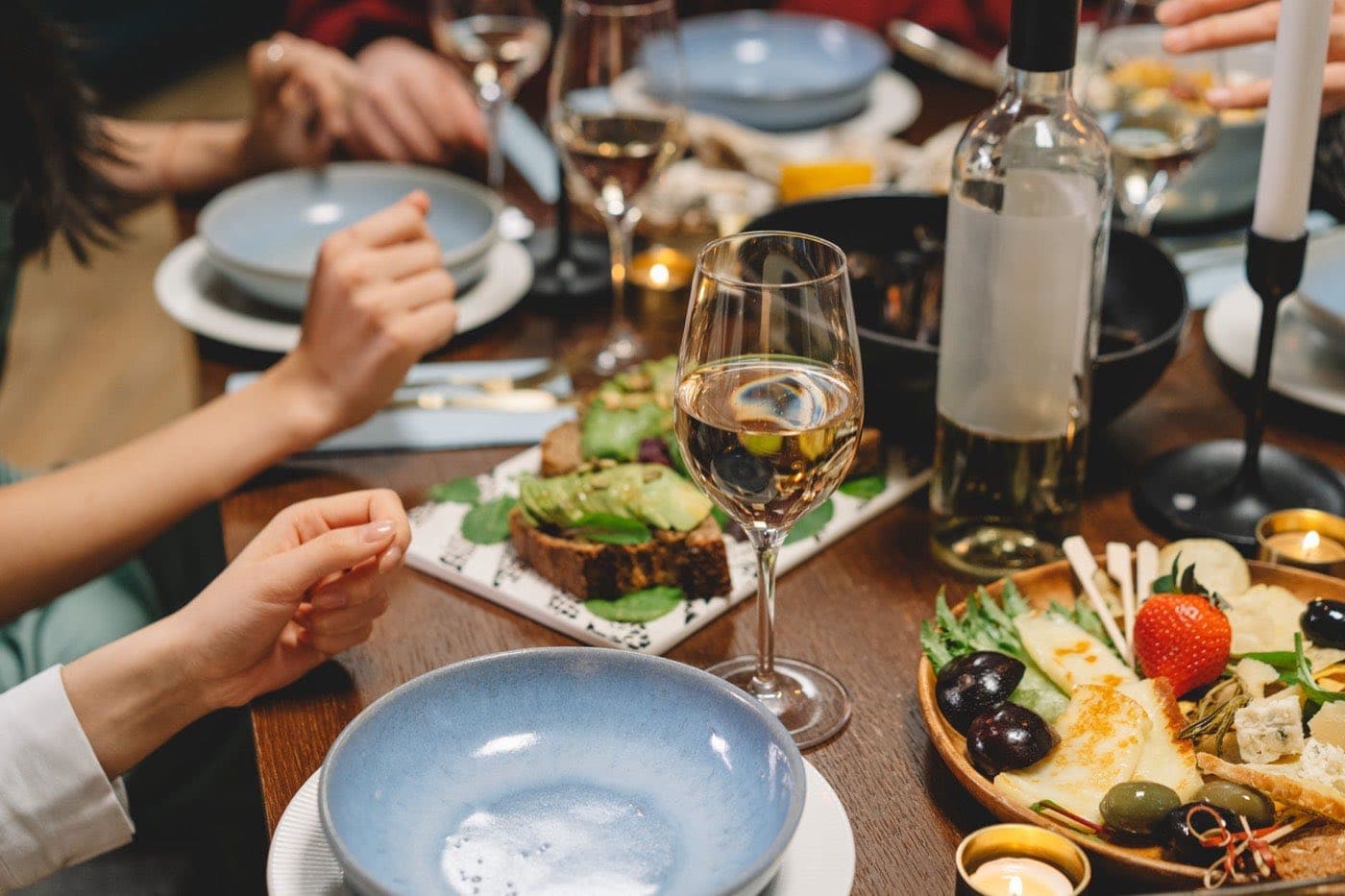 Friends sharing plates around a wooden table with white wine, a cheese board and avocado toast