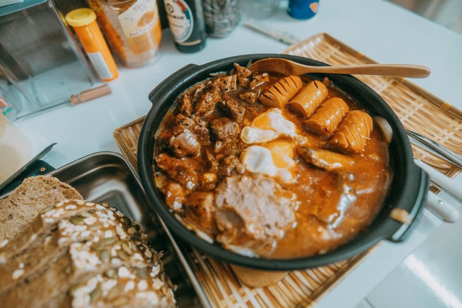 A pan of stew with braised beef, sausages and a soft egg in a red broth, next to a seeded loaf