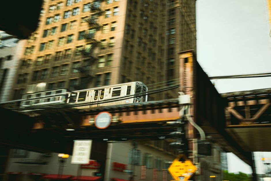A silver L train crossing a steel bridge above a street in the Chicago Loop, with a tall brick office building and its fire escapes behind it
