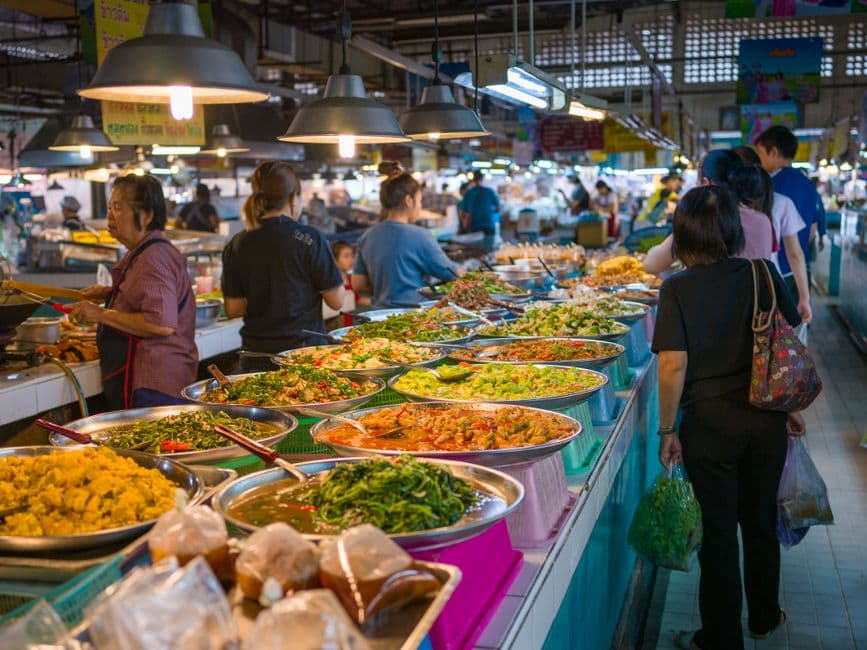 Trays of curries and stir-fried vegetables lined up at a covered Thai market food stall with customers queuing