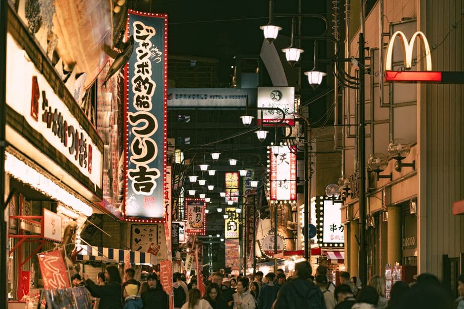 A crowded Osaka street at night lined with illuminated restaurant signs, lanterns and food stalls