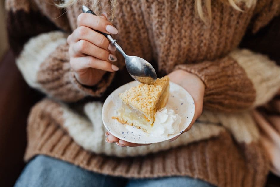 A person in a knitted sweater holding a small plate with a slice of crumb-topped apple pie and whipped cream