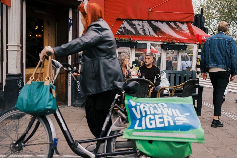 A woman wheeling a bicycle past a red cafe awning in Amsterdam while two people sit at the terrace behind her