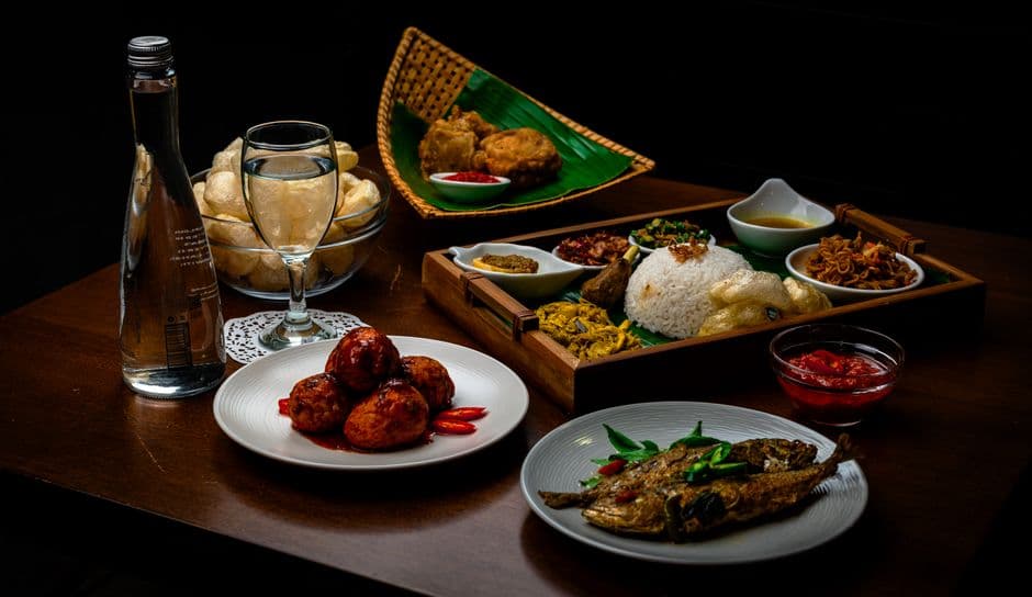 A wooden tray of rice ringed by small dishes of curry, sambal and fried shallots, with prawn crackers and a whole fried fish beside it
